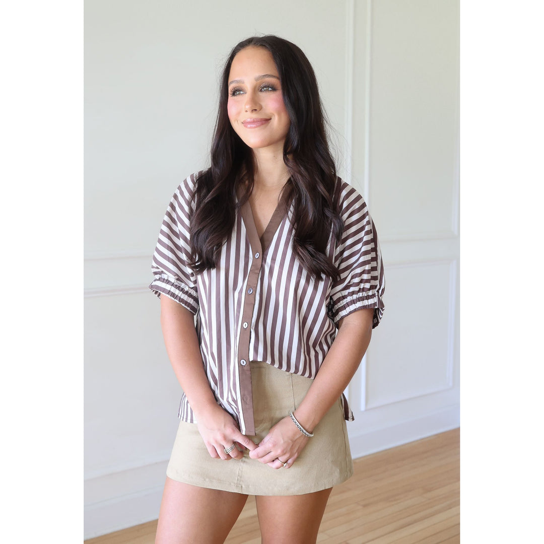 A woman modeling a brown and white vertically striped short-sleeve button-up shirt with ruffled sleeves, paired with a beige mini skirt, standing in a brightly lit room with white paneled walls and light wood floors.