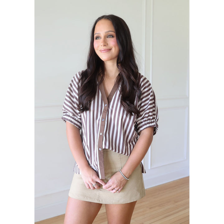A woman modeling a brown and white vertically striped short-sleeve button-up shirt with ruffled sleeves, paired with a beige mini skirt, standing in a brightly lit room with white paneled walls and light wood floors.