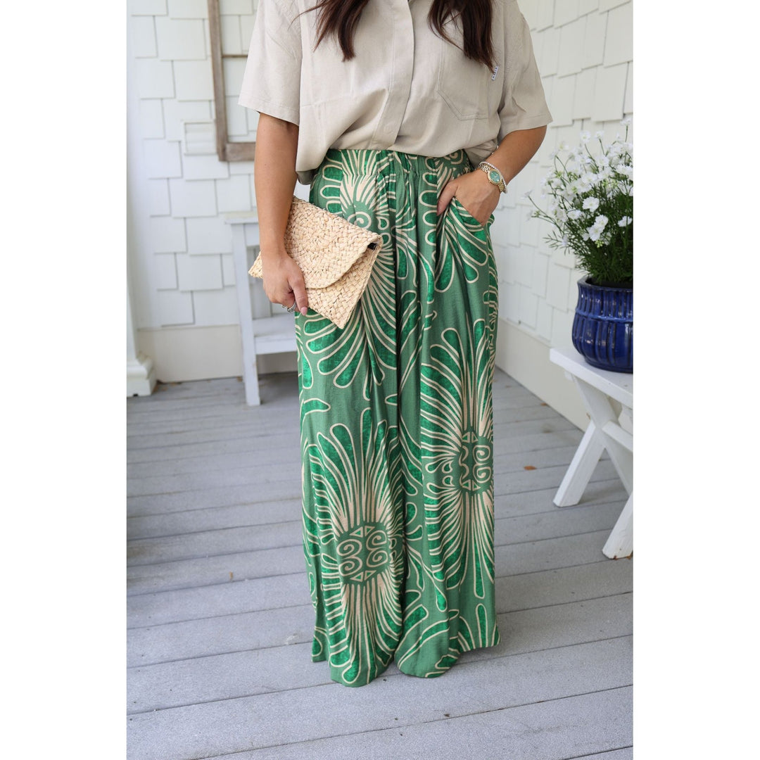 Person wearing green patterned pants with a beige top and clutch, standing on a wooden floor.