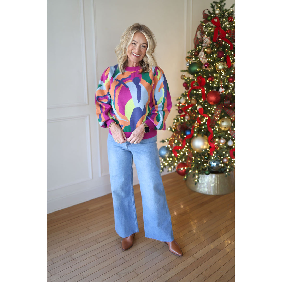 Woman wearing a colorful sweater and jeans standing in front of a decorated Christmas tree.