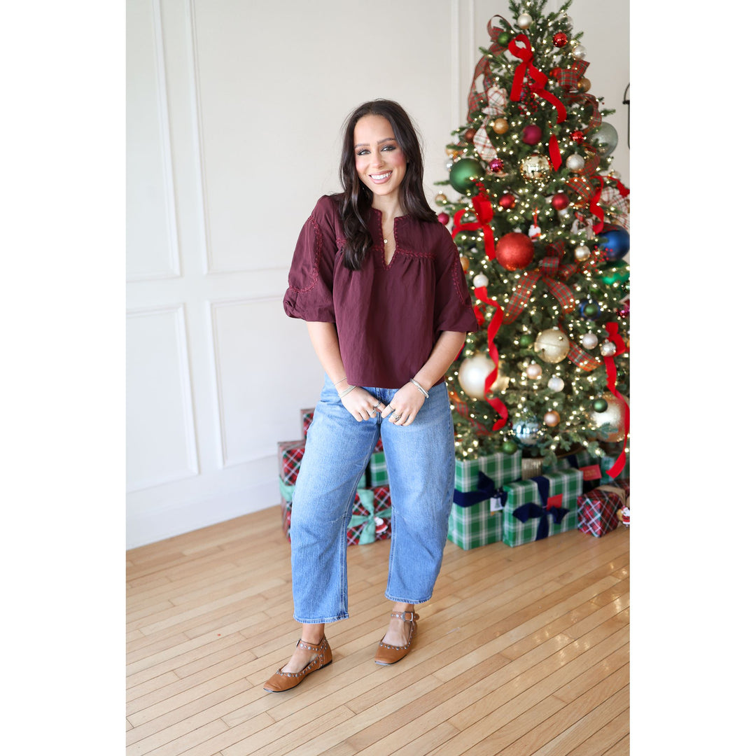 Woman standing in front of a decorated Christmas tree wearing a burgundy top and blue jeans.