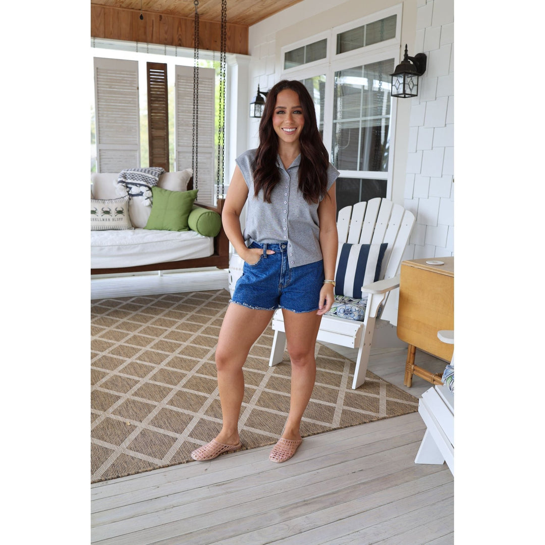 Woman standing on a porch wearing a gray sleeveless shirt and blue shorts.
