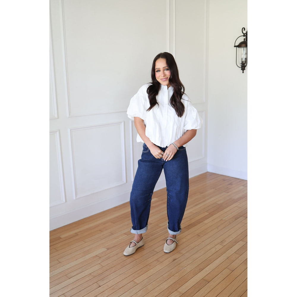 Woman wearing a white blouse and blue jeans standing in a room with wooden flooring and white walls.