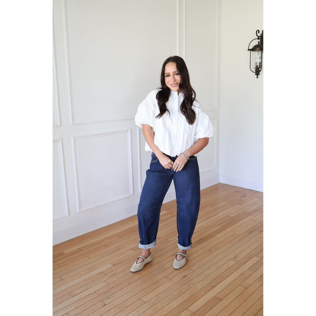 Woman wearing a white blouse and blue jeans standing in a room with wooden flooring and white walls.