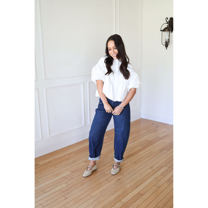 Woman wearing a white blouse and blue pants standing in a room with wooden flooring and white walls.