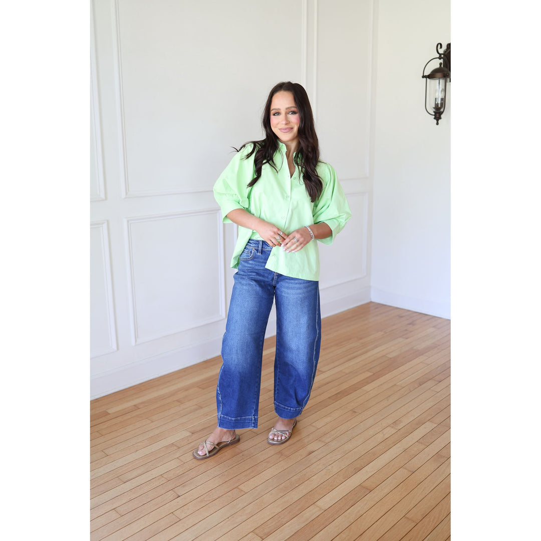 Woman wearing a bright green shirt and blue jeans standing in a room with wooden flooring and white walls.