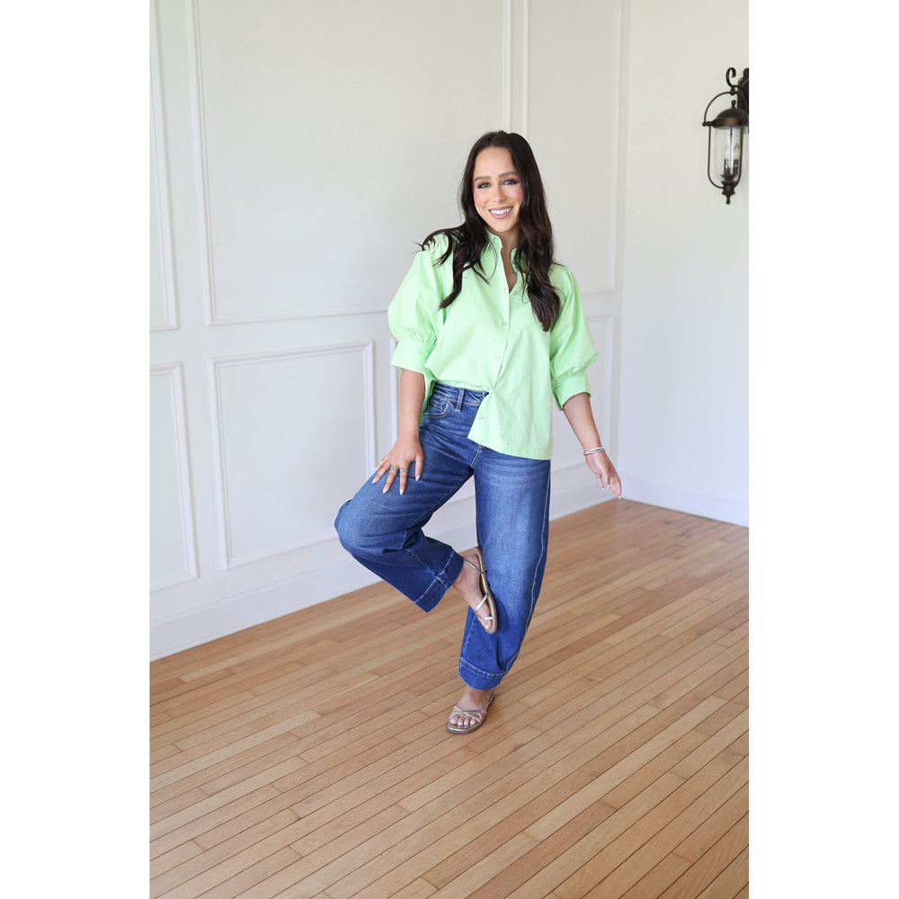 Woman wearing a bright green shirt and blue jeans standing in a room with wooden flooring and white walls.