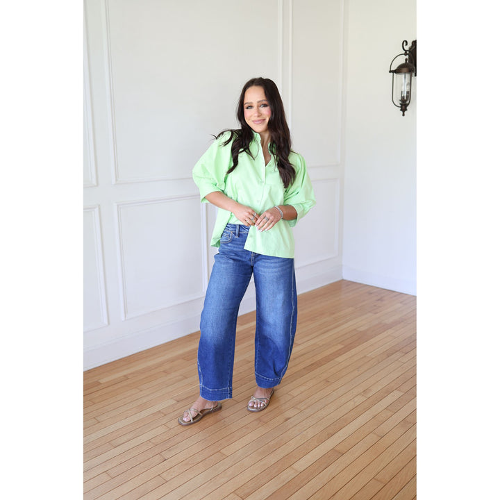 Woman wearing a bright green blouse and blue jeans standing in a room with wooden flooring and white walls.