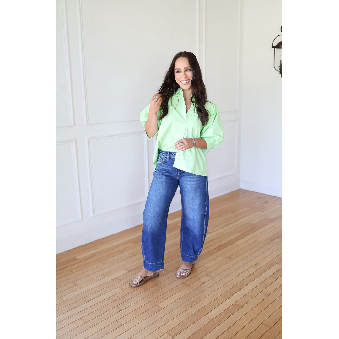 Woman wearing a bright green shirt and blue jeans standing in a room with wooden flooring and white walls.
