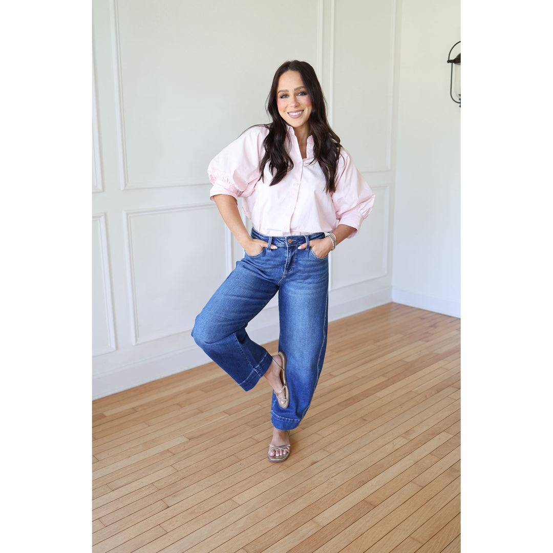 Woman wearing a pink blouse and blue jeans standing in a room with wooden flooring and white walls.