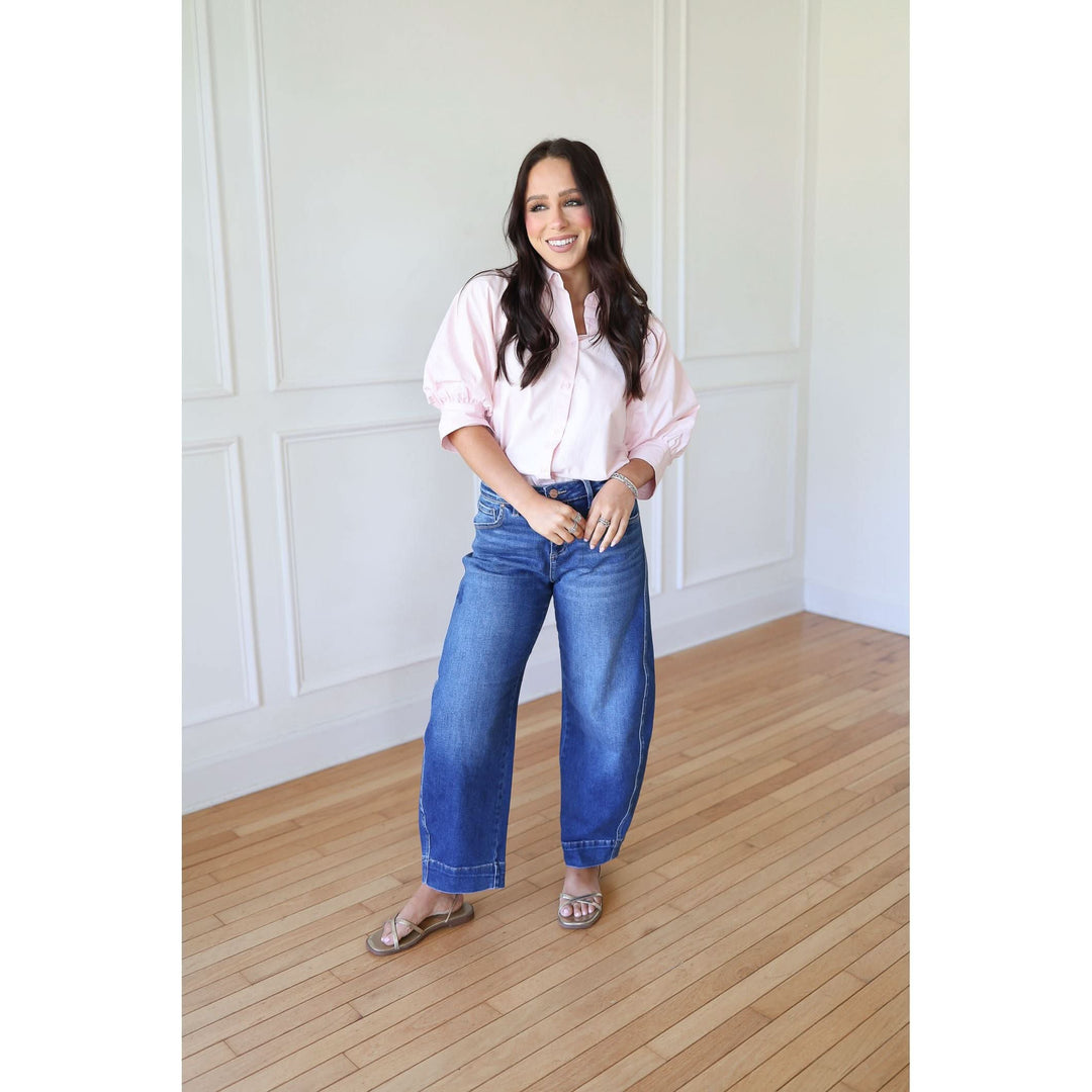 Woman wearing a light pink shirt and blue jeans standing in a room with wooden flooring and white walls.
