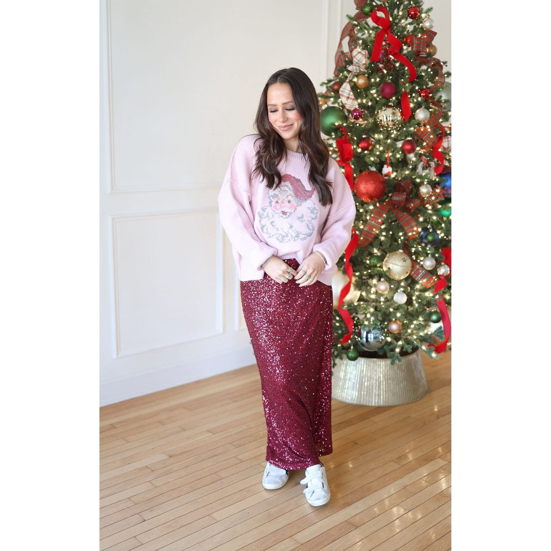 Woman wearing a pink sweater with a Santa face and a red sequin skirt standing in front of a decorated Christmas tree.