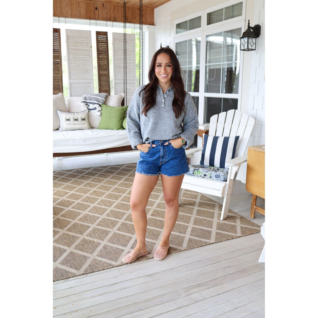 Woman standing on a porch wearing a gray sweater and blue shorts.
