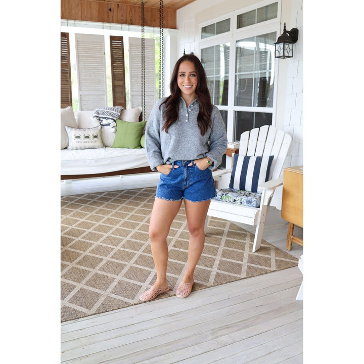 Woman standing on a porch wearing a gray sweater and blue shorts.