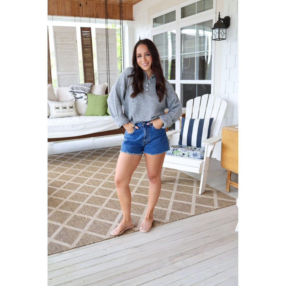 Woman standing on a porch wearing a gray sweater and blue shorts.