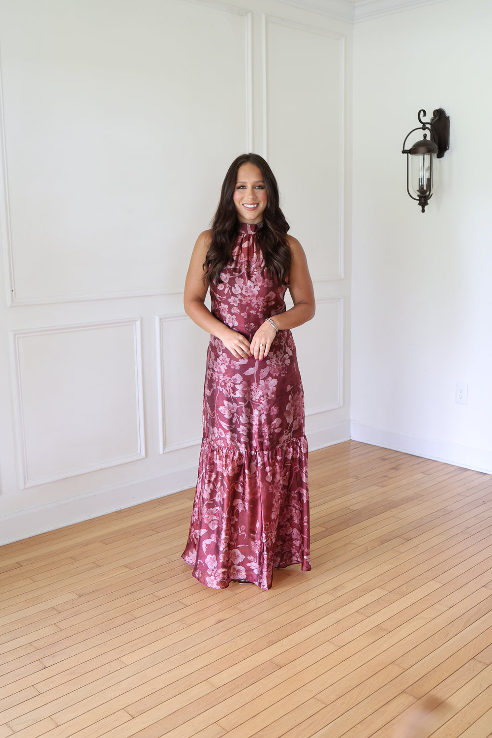 A woman wearing a sleeveless, high-neck, floor-length tiered gown with a pink and burgundy floral pattern