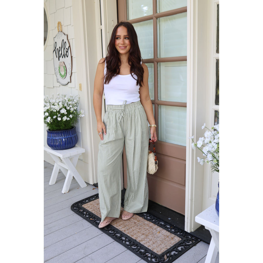 Woman standing on a porch wearing a white top and green striped pants.
