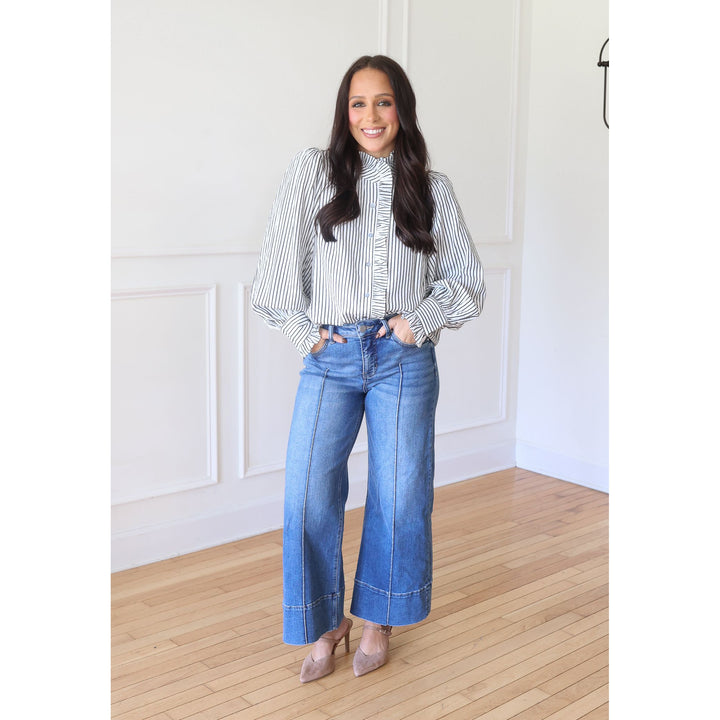 A woman stands in a bright room with white paneled walls and light wood floors, wearing a black and white vertically striped long-sleeve blouse with ruffled trim, blue wide-leg jeans, and nude pointed-toe heels.