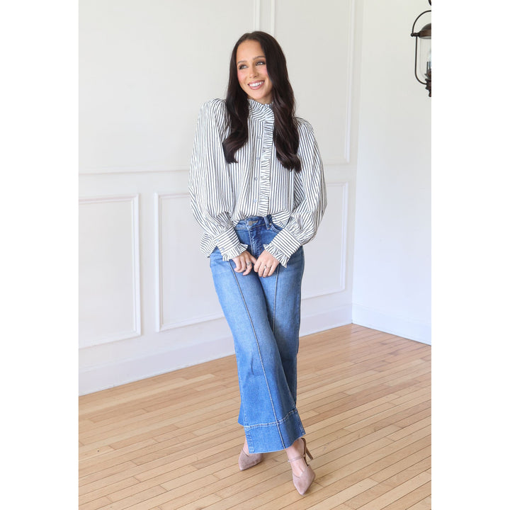 A woman stands in a bright room with white paneled walls and light wood floors, wearing a black and white vertically striped ruffled blouse, blue wide-leg jeans, and nude heels.