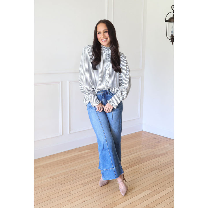 A woman stands in a bright room with white paneled walls and light wood floors, wearing a white long-sleeve blouse with thin black vertical stripes and ruffled trim, blue wide-leg jeans, and nude pointed-toe heels.