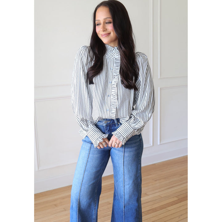A woman wearing a black and white vertically striped long-sleeve blouse with ruffle details at the collar and cuffs, paired with blue jeans, standing in a bright room with white paneled walls and light wood floors.