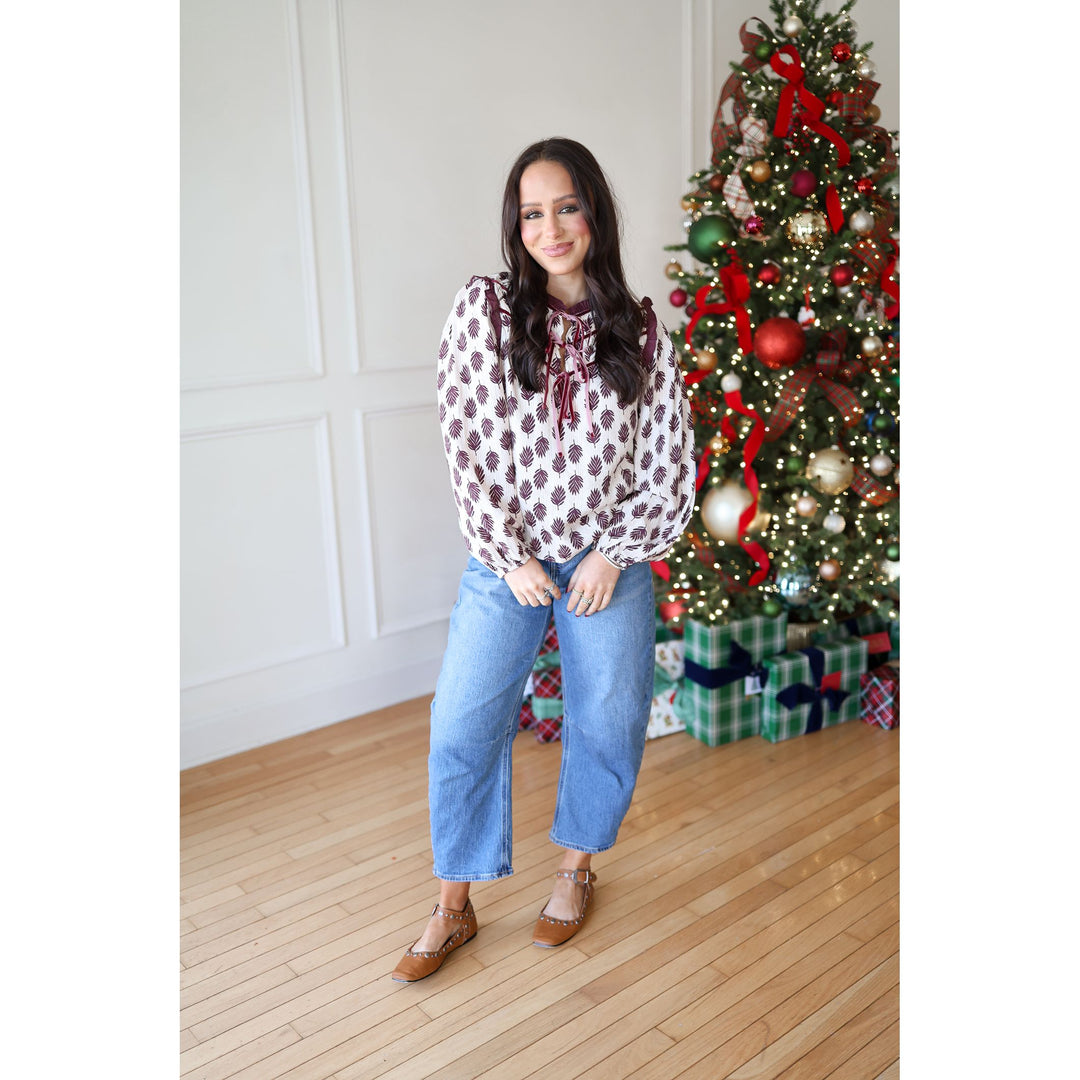 Woman standing in front of a decorated Christmas tree wearing a patterned blouse and jeans.