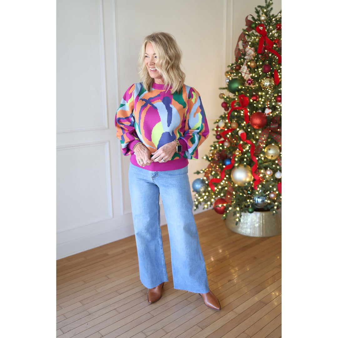 Woman wearing a colorful sweater and jeans standing next to a decorated Christmas tree.