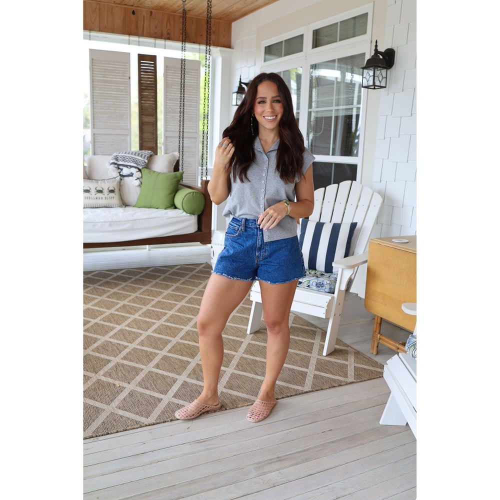 Woman standing on a porch wearing a gray shirt and blue shorts, smiling.