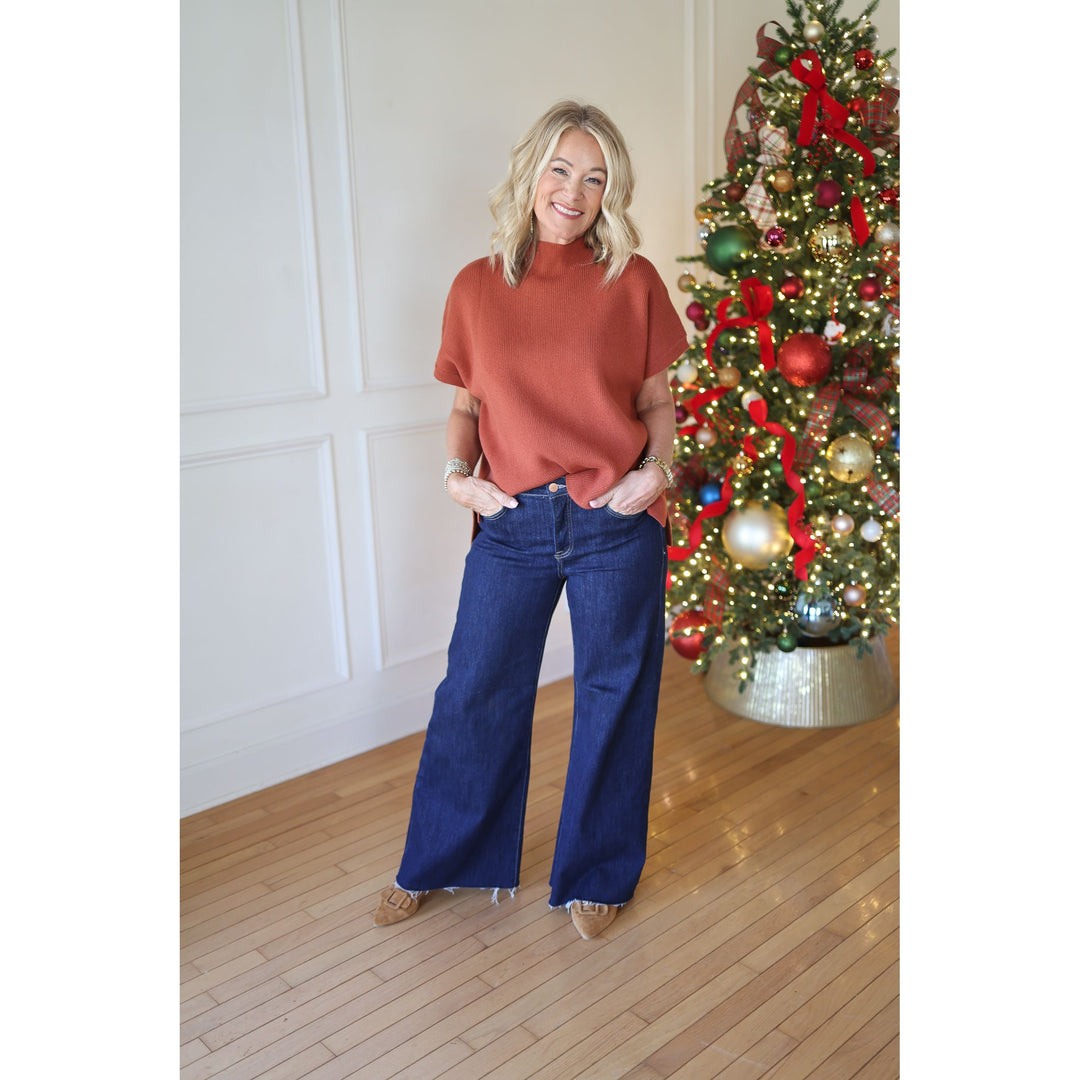Woman in a rust-colored sweater and blue jeans standing in front of a decorated Christmas tree.