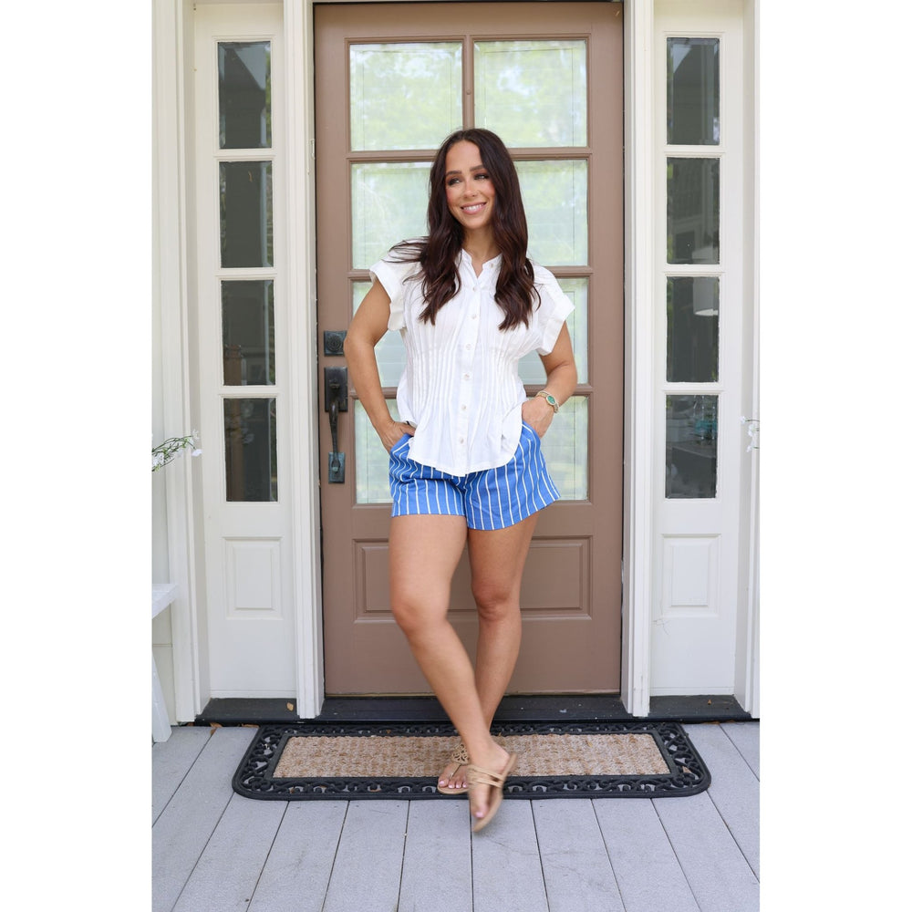 Woman standing in front of a brown door wearing a white blouse and blue striped shorts.