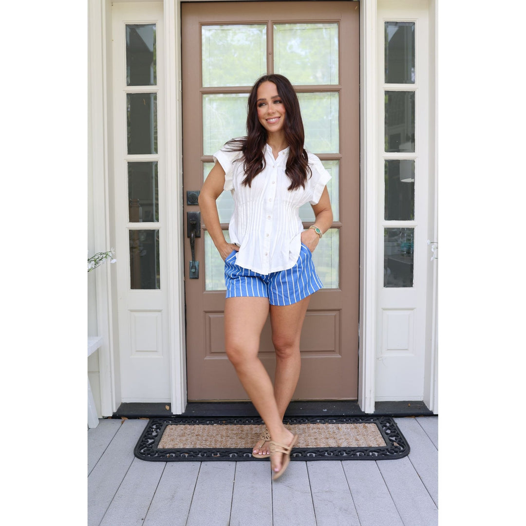 Woman standing in front of a brown door wearing a white blouse and blue striped shorts.