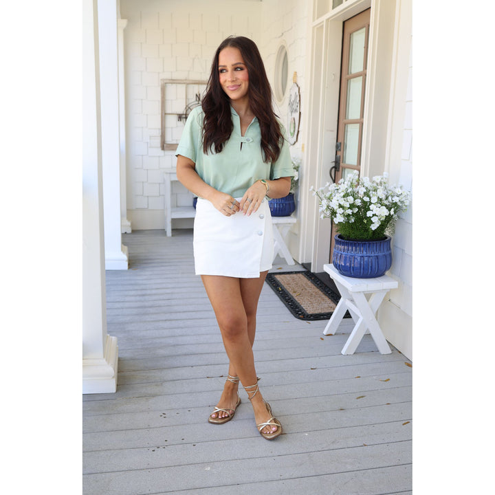 Woman in a light green top and white skirt standing on a wooden porch.