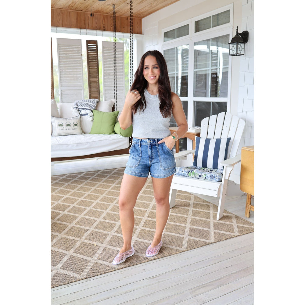 Woman standing on a porch wearing a gray tank top and denim shorts.