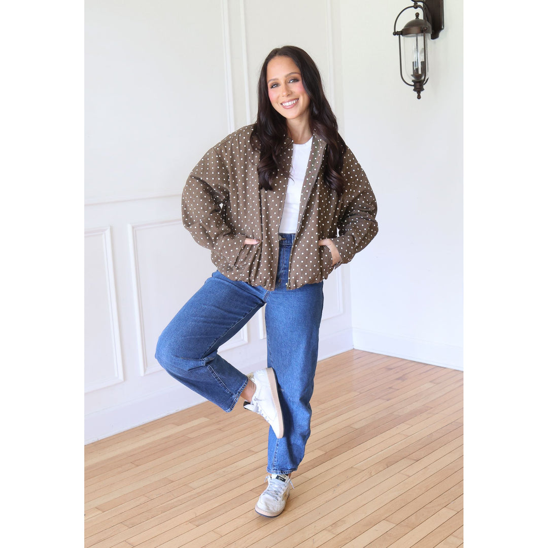 A woman posing in a brown and white polka dot bomber jacket, blue jeans, and white sneakers