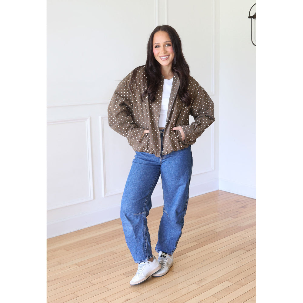 A woman stands wearing a brown bomber jacket with white polka dots, blue jeans, and white sneakers.