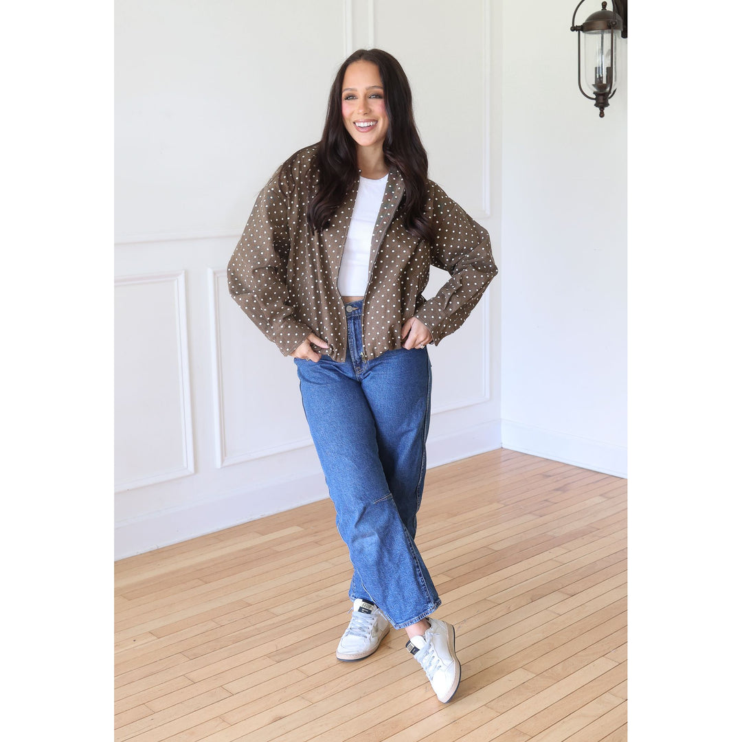 A woman stands wearing a brown long-sleeve button-up shirt with white polka dots, a white t-shirt, blue jeans, and white sneakers.