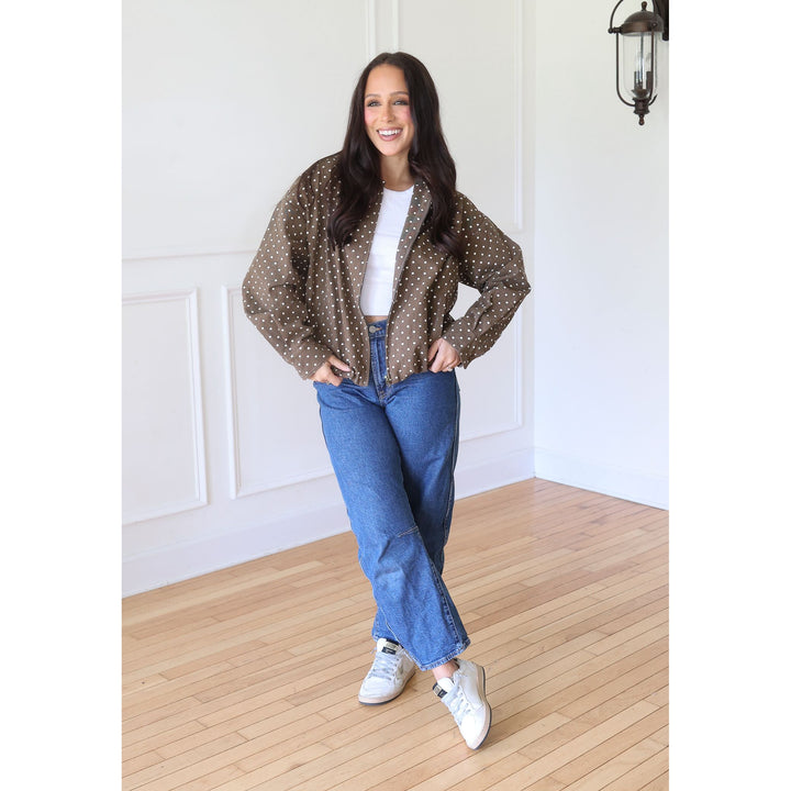 A woman stands wearing a brown long-sleeve button-up shirt with white polka dots, a white t-shirt, blue jeans, and white sneakers.