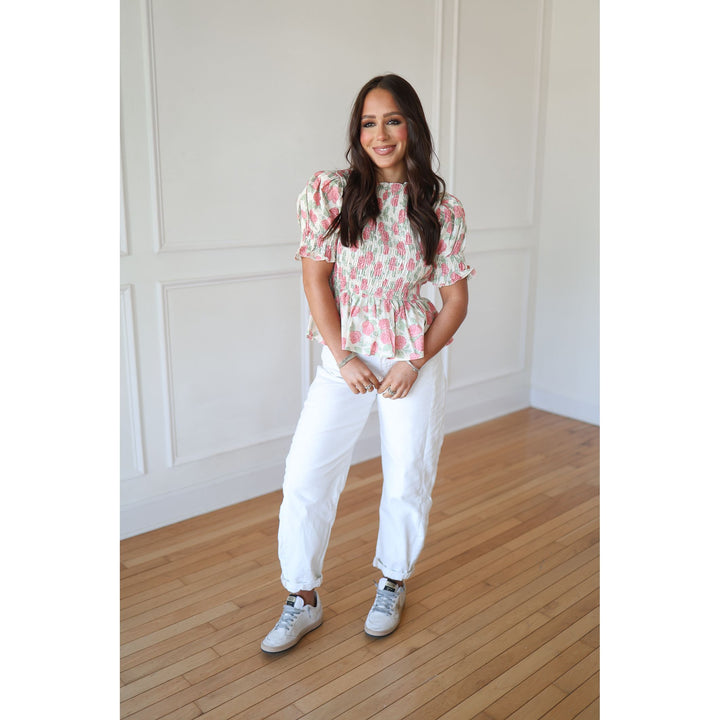 Woman wearing a floral blouse and white pants standing in a room with wooden flooring and white walls.