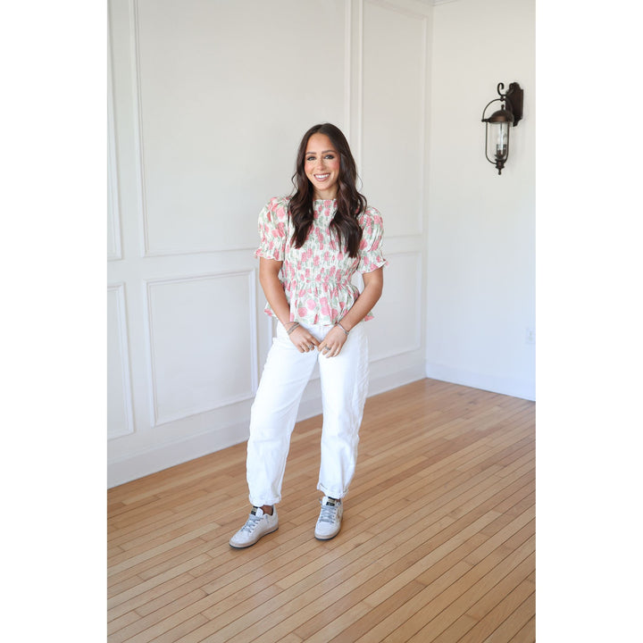 Woman in a floral blouse and white pants standing in a room with wooden flooring and white walls.