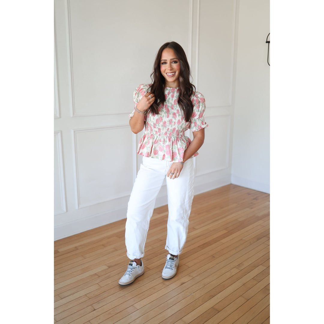 Woman wearing a floral top and white pants standing on a wooden floor.
