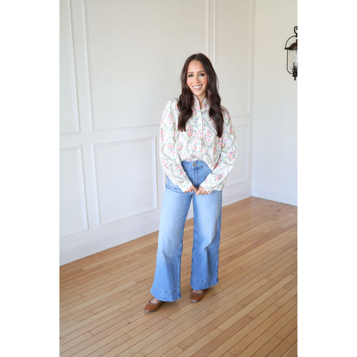 Woman wearing a floral blouse and blue jeans standing in a room with wooden flooring and white walls.