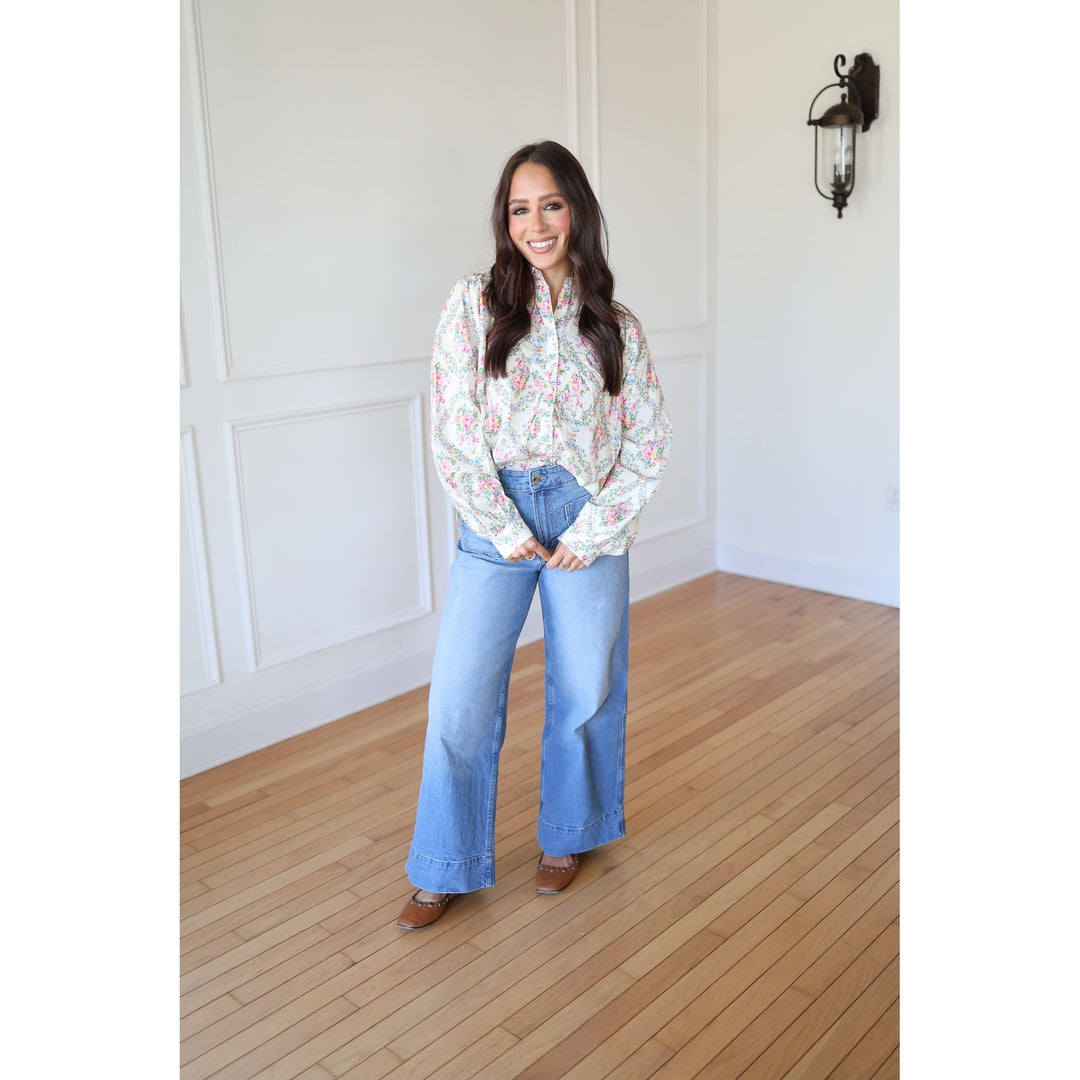 Woman wearing a floral blouse and blue jeans standing in a room with wooden flooring and white walls.