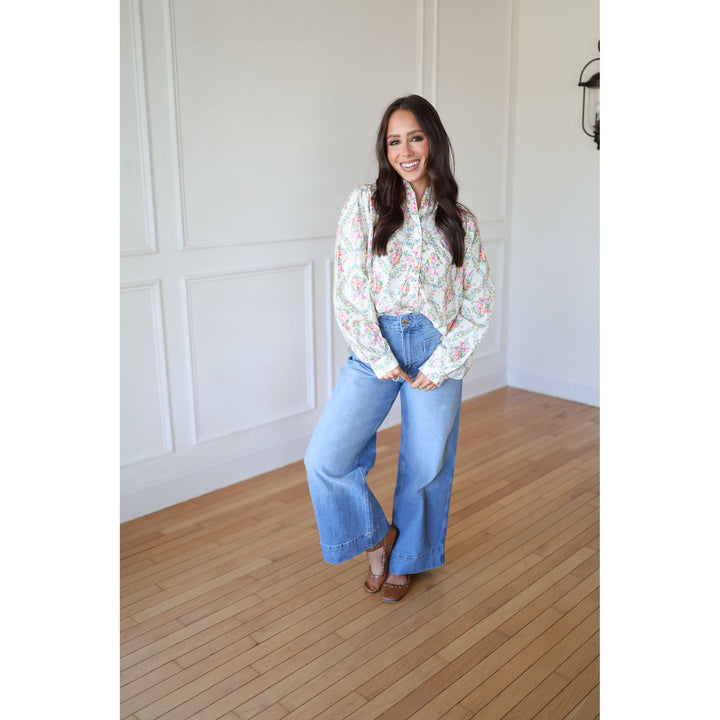 Woman wearing a floral blouse and blue jeans standing in a room with wooden flooring and white walls.