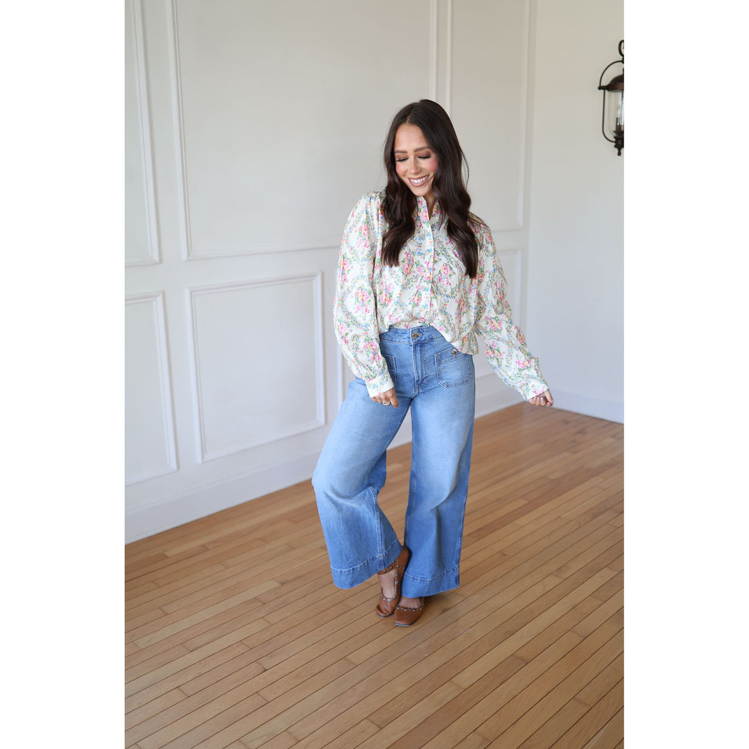 Woman wearing a floral blouse and blue jeans standing in a room with wooden flooring and white walls.