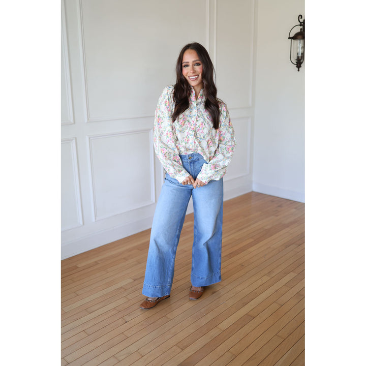 Woman wearing a floral blouse and blue jeans standing in a room with wooden flooring and white walls.