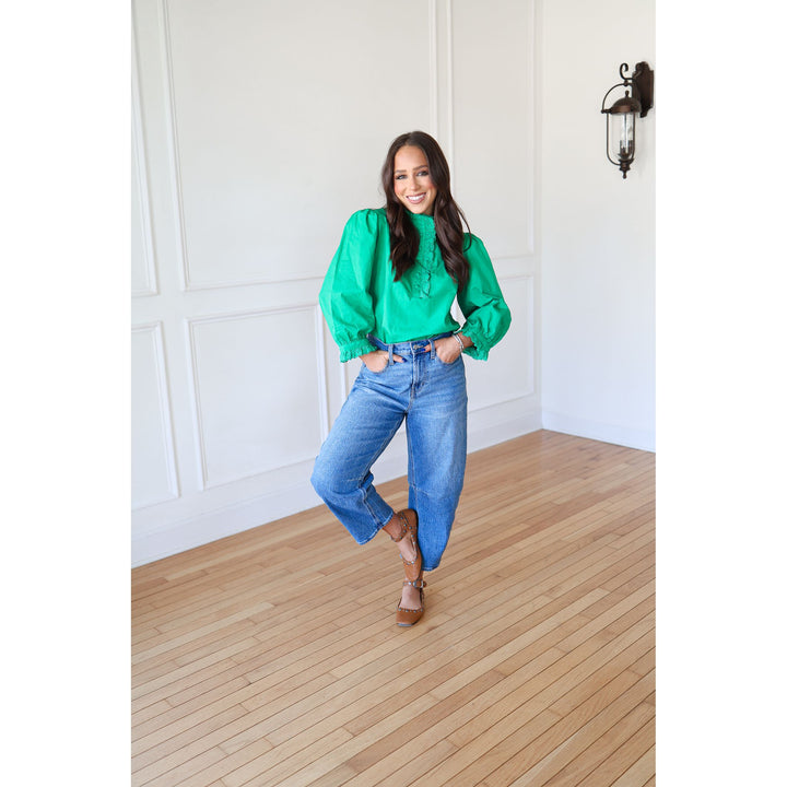 Woman in a green blouse and blue jeans standing in a room with wooden flooring and white walls.