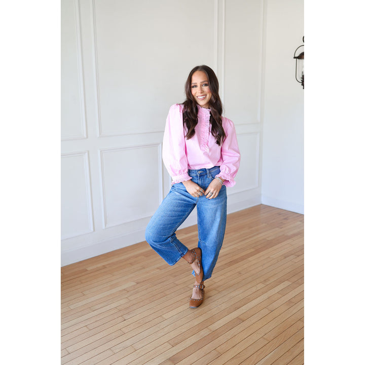 Woman in a pink blouse and blue jeans standing in a room with wooden flooring and white walls.