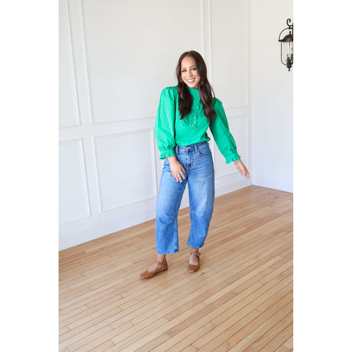 Woman wearing a green blouse and blue jeans standing in a room with wooden flooring and white walls.