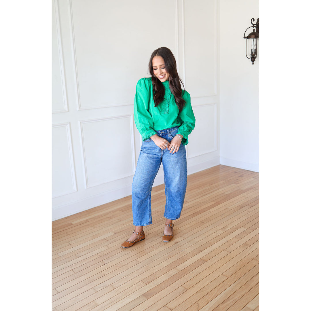 Woman in a green blouse and blue jeans standing in a room with wooden flooring and white walls.