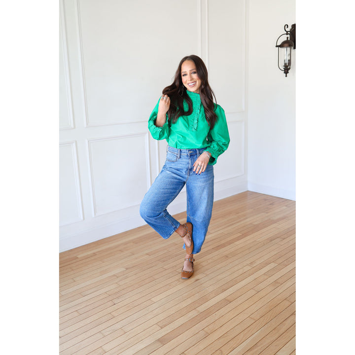 Woman in a green blouse and blue jeans standing in a room with wooden flooring and white walls.